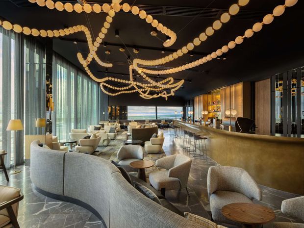 A sophisticated hospitality lounge or hotel bar with a long curved bar counter, extensive back-bar shelving, panoramic glazing, and multiple clusters of upholstered lounge seating. The dark ceiling is decorated with dramatic looping strands of warm globe lights, while large curtains and stone-look flooring create a refined, luxurious atmosphere. A distant view is visible through the glazing.