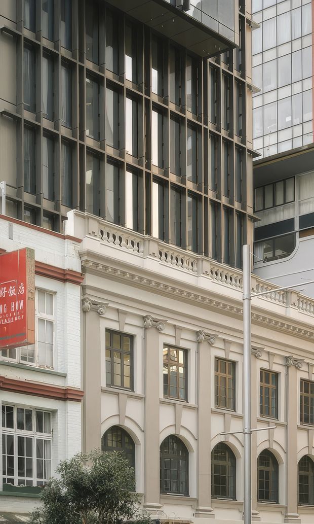 Street-level view of a dense urban building facade combining a preserved ornate heritage frontage with a contemporary multi-storey glazed addition above. The older lower facade has cream painted masonry, decorative cornices, pilasters, arched openings, timber-framed windows and a balustraded parapet. The upper extension uses dark metal framing, extensive vertical glazing and projecting horizontal slabs. Adjacent commercial frontage and signage are visible at the left.