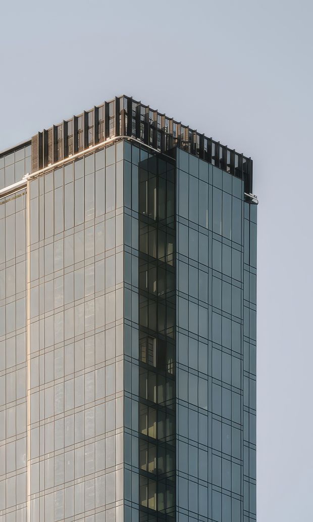 Close-up exterior view of the upper corner of a contemporary multi-storey commercial building. The facade is dominated by large blue-grey glazed curtain wall panels with dark metal mullions, while the roofline features a projecting parapet and repeated vertical dark metal fins or screens. The image focuses on the building envelope against a plain sky.