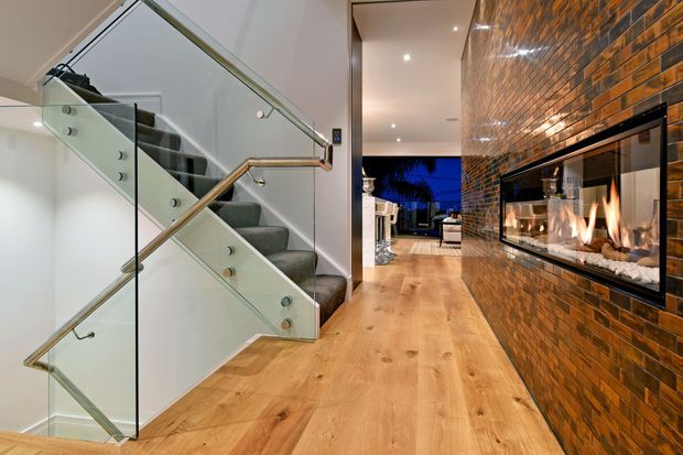 A contemporary residential hallway with warm timber flooring, a floating staircase enclosed by clear glass balustrades and timber handrails, and a full-height exposed brick feature wall incorporating a long linear gas fireplace. Recessed ceiling lights lead toward an open living area and exterior glazing at the rear.