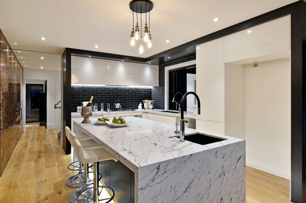 Contemporary kitchen with a large white marble island featuring an integrated sink and black tapware, white cabinetry, a glossy black subway-tile splashback, timber flooring, and black-framed architectural detailing. Pendant lights hang above the island, with bar stools providing seating.