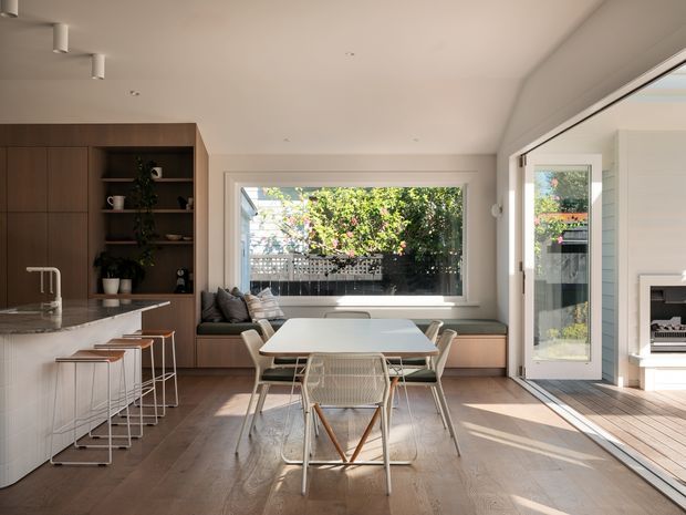 A bright, open-plan kitchen and dining area with warm timber cabinetry, a pale stone island, and a light timber floor. The dining table sits beside a large rear window and full-height glazed doors opening to an outdoor deck, creating strong indoor–outdoor connection. The room has clean contemporary lines, white walls and ceiling, integrated bench seating, understated pendant-free lighting, and a calm neutral palette with natural timber accents.