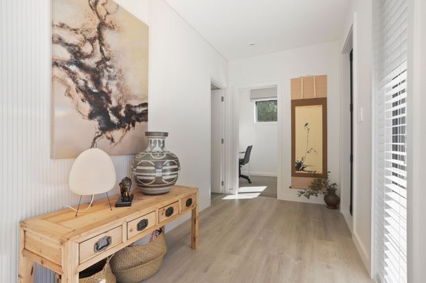 Bright, narrow residential hallway or entry passage with pale timber flooring, white vertically fluted wall panelling, and white plantation shutters. A rustic timber console with drawers and woven baskets sits beneath an abstract artwork, styled with a ceramic vessel and small lamp. The corridor leads toward a small home office or study area, with a tall timber-framed decorative niche visible at the far end.