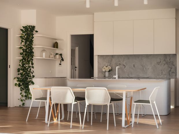 Bright open-plan kitchen and dining area with white flat-panel cabinetry, a large grey marble-look splashback, light stone island benchtop, and a white vertically tiled island face. A timber dining table with white chairs sits in the foreground, alongside built-in open shelving and a tall indoor plant. The room has warm timber flooring, soft neutral finishes, and simple ceiling track lighting.