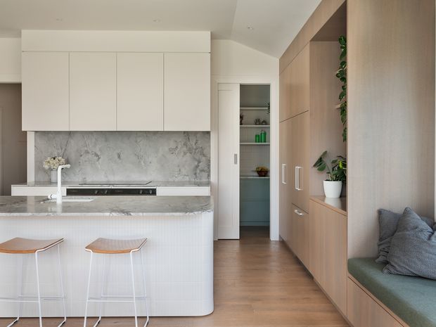 Bright contemporary kitchen with a white marble-look splashback and benchtop, integrated white cabinetry, a curved island with fluted detailing and two timber-topped stools. Full-height warm timber cabinetry lines the right wall and incorporates a built-in upholstered window seat. A compact pantry or storage room is visible through the doorway at the rear.