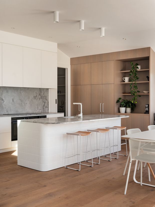 Bright contemporary kitchen and dining area with a large white island clad in small vertical tiles, a pale stone benchtop, white handleless overhead cabinetry, marble-look splashback, warm timber flooring, and a full-height timber storage wall with open shelving and plants. White stools line the island, with a dining table and chairs visible at right.