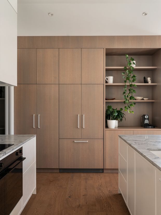 A refined contemporary kitchen with a warm timber wall of integrated cabinetry and open display shelving. Two pale island benches with veined stone tops frame the view, while dark timber flooring, recessed ceiling lights, stainless handles, appliances, ceramics, and trailing plants complete the space.