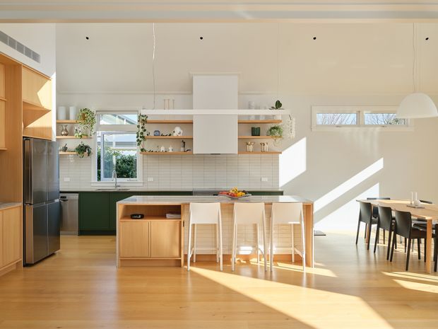Bright open-plan kitchen and dining area with warm timber flooring and cabinetry, a dark green kitchen run, white tiled splashback, open timber shelving, and a central island with pale seating. Large windows and high ceilings bring in strong natural light, while a dining table sits to the right.