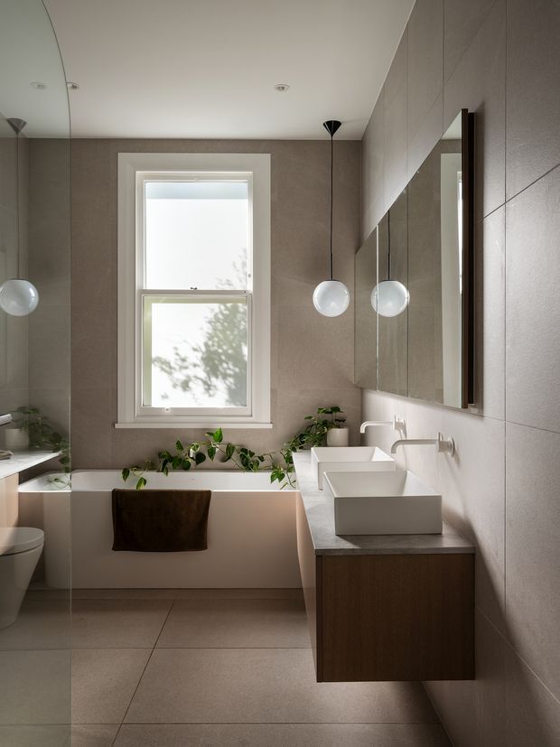 A refined contemporary bathroom with a freestanding-style rectangular bathtub beneath a window, a wall-mounted dark timber vanity with two white vessel basins, large mirrored cabinetry, and twin globe pendant lights. Large-format taupe-grey tiles cover the walls and floor, while indoor plants add a natural accent. A toilet is partially visible at left.