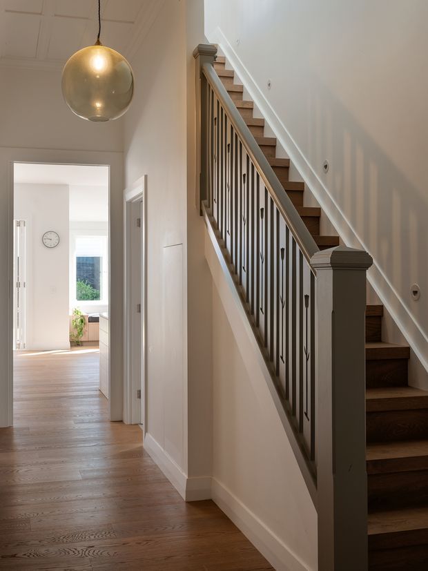 A bright residential hallway with a timber staircase rising along the right wall. The staircase features dark stained timber treads, painted white stringers and posts, and slender dark metal balusters with decorative details. Warm timber flooring continues through the corridor, with white paneled walls, simple trim, and a large spherical pendant light. Open doorways lead to a sunlit room in the background.