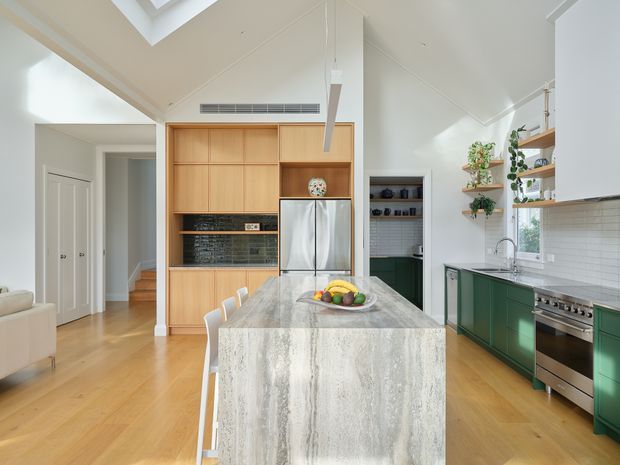 Bright, airy kitchen with a large waterfall-edge stone island, green base cabinetry, pale timber wall cabinetry, stainless-steel appliances, white tiled splashback, and warm timber flooring. The kitchen has a vaulted ceiling with skylights, a linear pendant light, open shelving with plants, and an adjacent hallway and stairway visible in the background.