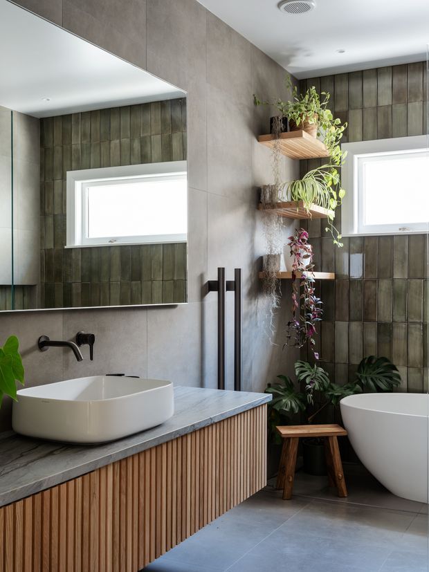 A contemporary bathroom featuring a floating timber slat vanity with a grey stone benchtop, white vessel basin, matte black wall-mounted tapware, and a freestanding white bathtub. Large mirrors, grey wall panels, vertically patterned dark tiles, open timber plant shelves, and abundant greenery create a warm, natural spa-like setting.
