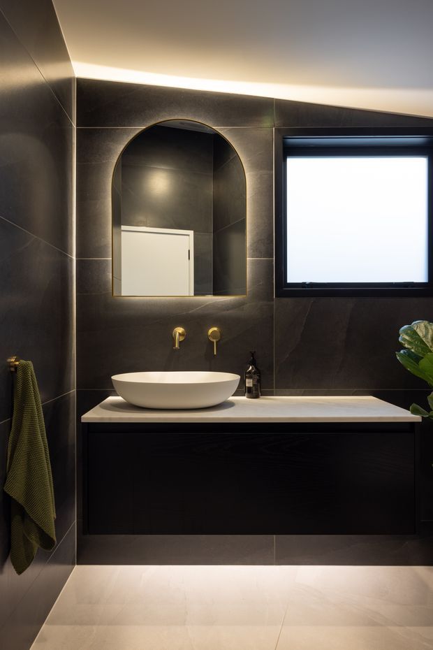 A moody contemporary bathroom featuring large dark stone-look wall tiles, a floating dark timber vanity with a pale stone benchtop, a white vessel basin, wall-mounted brass tapware, an arched illuminated mirror, and a black-framed frosted window. Warm concealed lighting and a muted green towel add contrast.