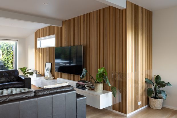 Contemporary living room with dark leather sofas, a wall-mounted television, white floating media cabinetry, indoor plants, and a full-height vertical timber slat feature wall wrapping around the room. Natural light enters through the windows, creating a warm and refined interior.