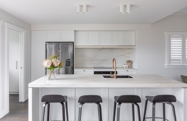 Bright, spacious contemporary kitchen with white Shaker-style cabinetry, a large pale stone island with integrated sink and brushed copper tap, four dark upholstered bar stools, stainless-steel refrigerator, and a veined stone backsplash. White plantation shutters and light neutral finishes create a clean, refined look.
