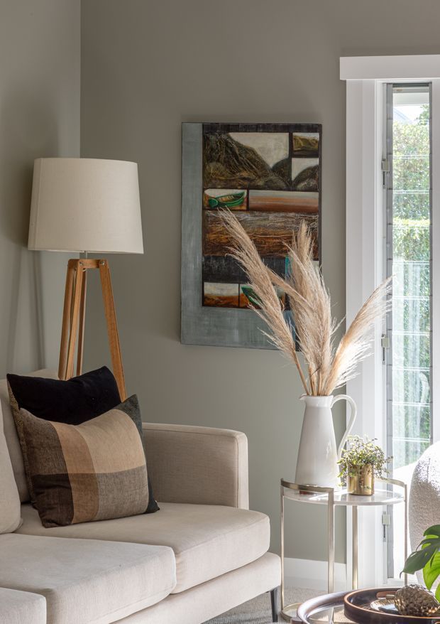 A softly styled living room corner featuring a neutral upholstered sofa with textured earth-toned cushions, a timber tripod floor lamp, abstract wall artwork, and a small round side table with a white vase of dried grasses. Tall glazed doors or windows bring in natural light, while muted taupe-grey walls and warm beige furnishings create a calm, organic atmosphere.