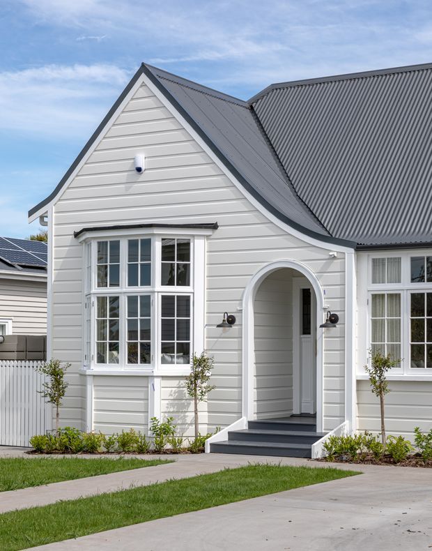 Exterior view of a light-grey, gable-roofed house with horizontal weatherboard cladding, white trim, a prominent arched entry porch, multi-pane bay window, dark metal roofing, paved paths, and restrained front landscaping.