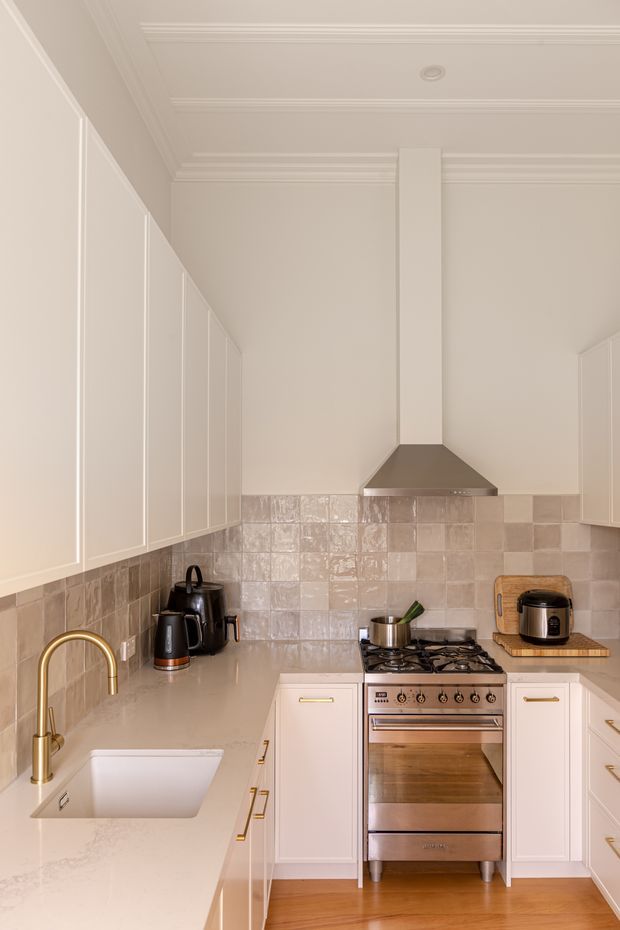 A compact, refined kitchen with white shaker-style cabinetry, pale stone benchtops, a glossy beige tiled splashback, and warm timber flooring. The room features a freestanding stainless-steel range cooker with a matching metal rangehood, a white undermount sink, and a brass gooseneck tap. Decorative ceiling mouldings and a central structural column add traditional architectural detailing, while small appliances sit on the benchtops.