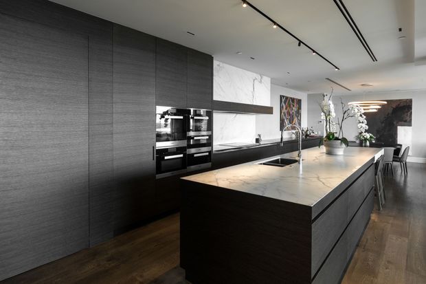 A spacious contemporary kitchen with a long dark textured built-in cabinetry wall, integrated double ovens, a large pale stone island with an undermount sink and gooseneck tap, and a dark stone or composite benchtop along the rear counter. Track lighting and linear ceiling details add a refined modern feel. Timber flooring, dining seating, artwork, and decorative plants are visible in the background.