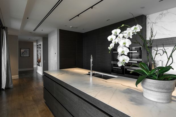 A sophisticated contemporary kitchen centered on a large dark-wood island with a pale veined stone benchtop, integrated sink, and minimalist tapware. Full-height dark cabinetry forms the rear wall, with open shelving and a dramatic white marble-look feature surface. Track lighting and recessed ceiling lights provide clean illumination, while an orchid adds a natural decorative element. Dark timber flooring continues into the adjacent hallway.