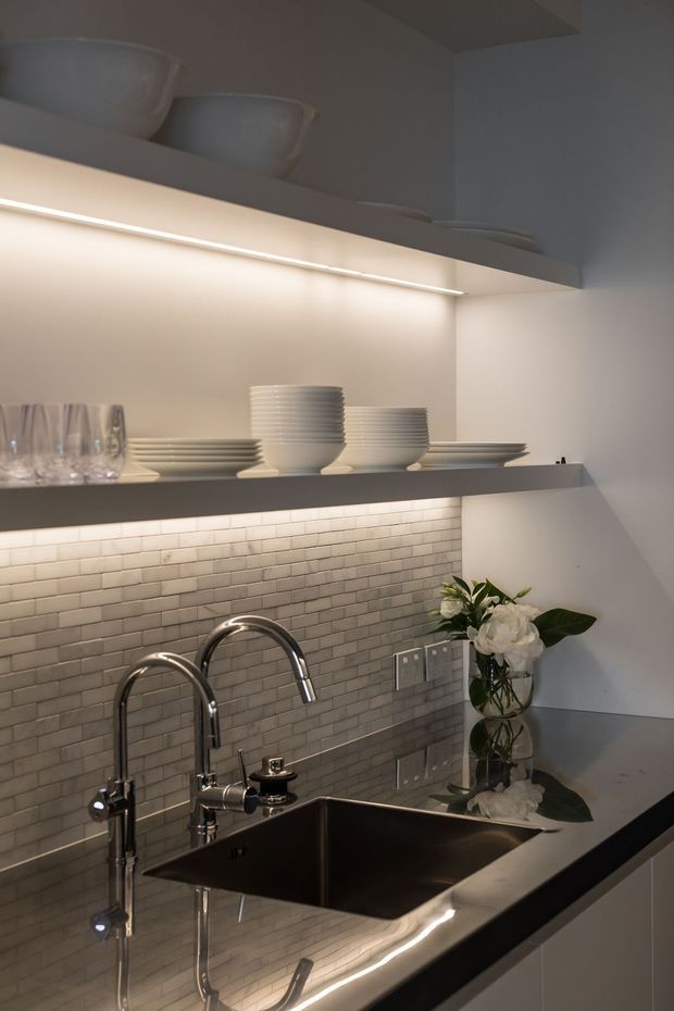 Close-up of a refined kitchen wet area with a deep stainless-steel sink, chrome gooseneck and secondary taps, glossy dark benchtop, and a grey brick-pattern tiled splashback. Illuminated open shelving displays neatly stacked white crockery and glassware, with a small floral arrangement adding a soft accent.