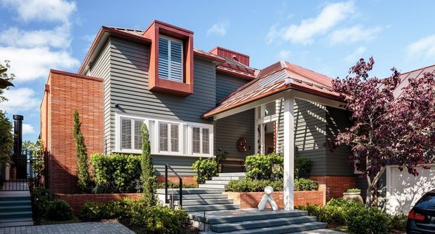 Exterior view of a multi-level suburban residence with dark grey horizontal weatherboard cladding, red brick sections, white plantation-shuttered windows, and a prominent red metal roof over the covered entry porch. The front garden includes clipped hedges, shrubs, paving and entry steps.