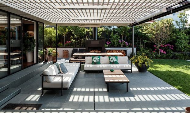 A contemporary outdoor living area beneath a large pergola with a translucent or slatted roof structure. The space features grey tiled paving, white upholstered sofas with green patterned cushions, a low timber coffee table, integrated outdoor dining and barbecue cabinetry, dark-framed glazing and steps to the house, and a landscaped garden beyond. Strong linear shadows create a bright, structured atmosphere.