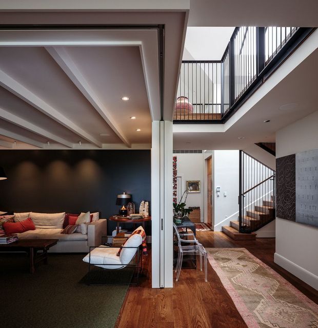 Interior view of a contemporary multi-level home combining a living room and open circulation area around a double-height stair void. The living room has a dark feature wall, sectional sofa, armchair, and layered lighting, while the adjacent hallway features timber flooring, a runner rug, artwork, and a staircase with slender black metal balustrades. An upper-level bridge and open stairwell create a strong vertical connection between floors.