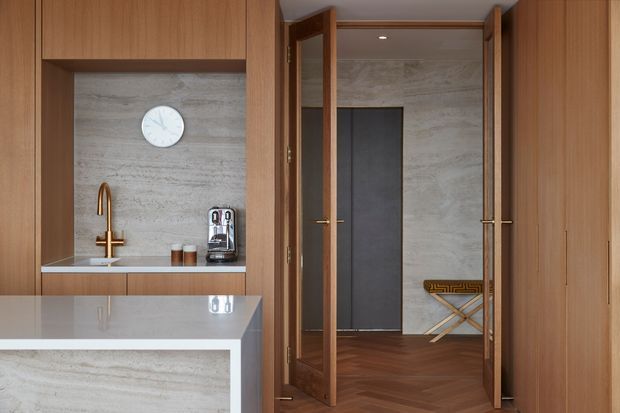 A refined residential interior showing a compact kitchen or beverage station opening through timber-framed double doors to a hallway or adjoining space. The built-in cabinetry is warm mid-tone timber, with a pale grey stone or marble-look wall finish and benchtop. A brass-toned tap, coffee machine, clock, and small accessories sit within the cabinetry, while the adjoining area has dark herringbone timber flooring, grey wall surfaces, and a folding stool. The composition is clean, tailored, and sophisticated with warm natural materials.