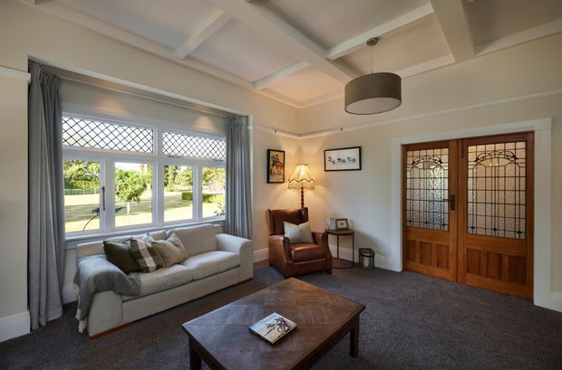 A traditional living room with a coffered ceiling, pale walls, a large leaded-glass window, and timber double doors with decorative glazing. The room contains a light upholstered sofa, brown leather armchair, timber coffee table, side table, framed artwork, and layered curtains. The restrained neutral palette and classic architectural detailing create a warm, refined atmosphere.