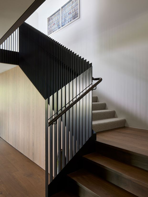 Interior view of a modern stairway with dark timber treads and flooring, a carpeted upper flight, black vertical metal balusters, and a dark timber handrail. Pale timber wall panelling encloses the stair structure, while white walls and a high window create a bright, double-height-feeling stair hall.