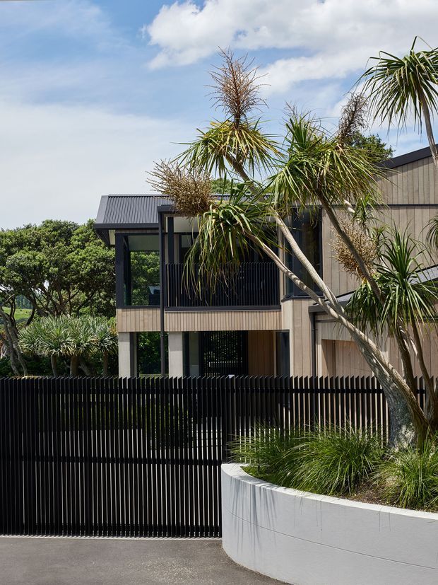 Street-facing view of a contemporary multi-level residence with a dark corrugated metal roof, vertical timber-toned facade cladding, extensive glazing, and a black vertical-slat fence and gate. A curved white rendered planter wall with spiky coastal landscaping frames the foreground, while mature trees and a bright sky form the background.