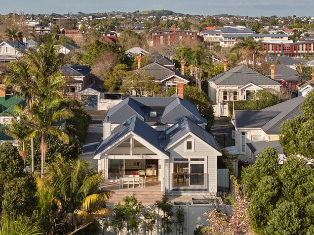 Elevated view across a leafy suburban neighbourhood, with a prominent multi-gabled light-coloured house in the foreground. The home features a steep pitched roof, prominent front gable, covered upper-level balcony or veranda, timber-style detailing, and a landscaped garden with mature palms. Numerous similarly pitched-roof houses and larger institutional or commercial buildings are visible in the background.