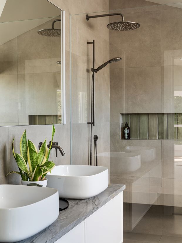 Contemporary bathroom with a walk-in glass shower, dual white vessel basins on a grey marble-look vanity, large-format beige-grey wall and floor tiles, dark bronze or black shower fittings, recessed shower niche, and a large wall mirror. A potted snake plant adds greenery to the otherwise neutral, refined space.