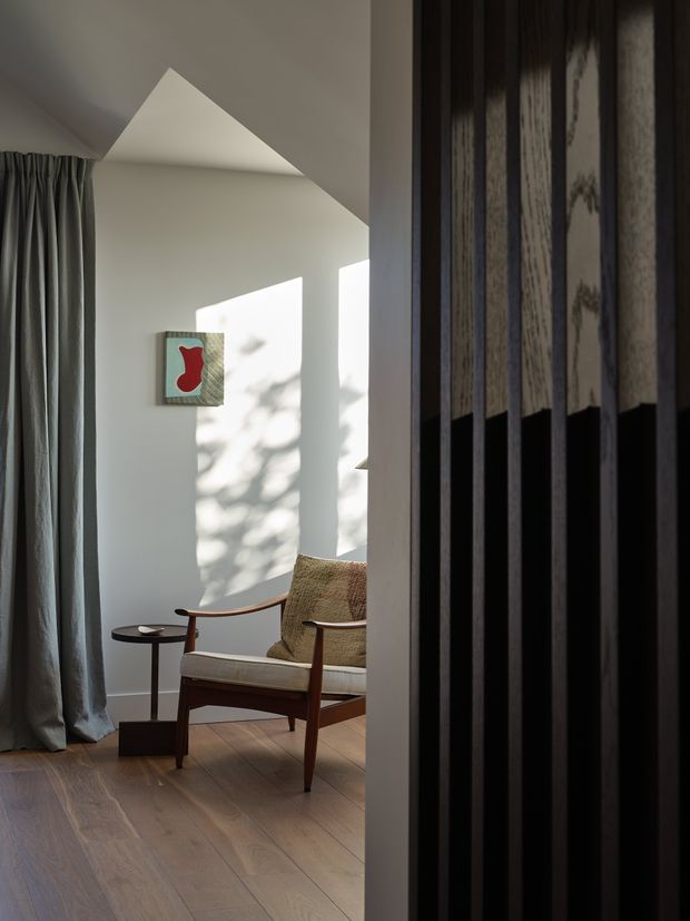 A softly lit contemporary sitting area viewed through a dark vertical timber screen. The room features pale walls, a warm timber floor, full-height grey curtains, and a mid-century style wooden lounge chair with woven upholstery beside a small dark side table. Strong angular sunlight and tree shadows fall across the wall, while an abstract artwork adds a restrained red accent. The ceiling includes a sharply angled architectural form.