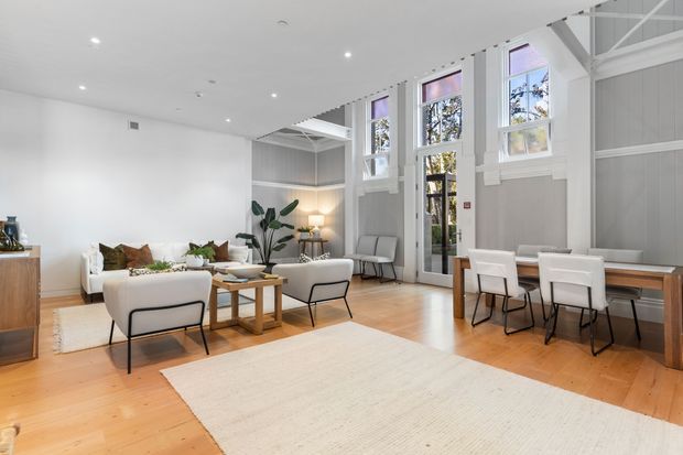 Bright open-plan living and dining room with white walls, light timber flooring, tall multi-pane windows and glazed doors, exposed white roof trusses, and high ceilings. The room contains a neutral sofa arrangement, timber coffee tables, a dining table with white chairs, rugs, indoor plants, and simple contemporary furnishings. Grey vertical wall panelling and abundant daylight create an airy, refined atmosphere.