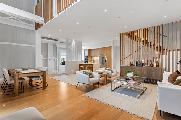 Bright, open-plan contemporary interior combining living, dining, and kitchen zones. The room features warm timber flooring, white walls and ceiling, pale upholstered seating, a timber dining table, a glass-and-metal coffee table, integrated timber kitchen cabinetry, and prominent vertical timber slat screens dividing the living area from the kitchen and circulation spaces. A partial upper-level void or stair balustrade is visible, contributing to the spacious feel.