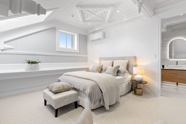 Bright, refined bedroom with a large upholstered bed, layered neutral bedding, a matching footstool, bedside table and lamp, and extensive white decorative wall and ceiling moulding. The room features white vertical wall panelling, a small window, ornate pressed ceiling details, and an arched opening into a bathroom with a timber vanity visible beyond.
