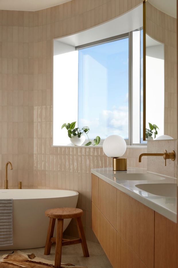 A warm, contemporary bathroom with a freestanding white bathtub, wall-mounted double vanity, large window, and mirrored cabinet. Glossy pale beige vertically arranged wall tiles curve around the room, while the vanity uses light timber cabinetry and a pale stone benchtop. Brushed brass tapware and a spherical lamp add refined accents, with a small wooden stool and plants on the windowsill.