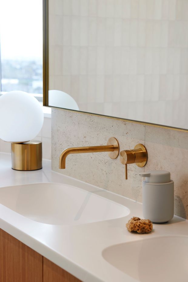 Close-up of a refined contemporary bathroom vanity with a white integrated double basin, wall-mounted brushed gold tapware, pale textured stone backsplash, large gold-edged mirror, and warm timber cabinetry. A sculptural globe lamp and minimal soap dispenser add soft decorative accents.