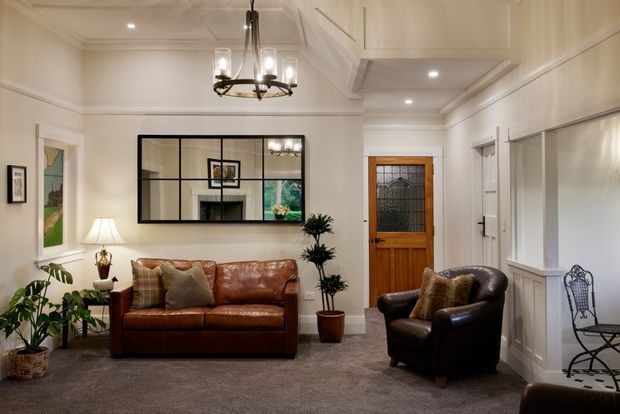 A refined traditional living room with cream wall panelling, decorative ceiling detailing, recessed lighting, and a dark metal chandelier. The room contains a brown leather sofa and armchair, a large black-framed internal mirror or glazed partition, potted plants, framed artwork, a timber-and-glass entry door, and grey carpet. Built-in white cabinetry or panelling lines the right wall, creating a warm, classic sitting area.