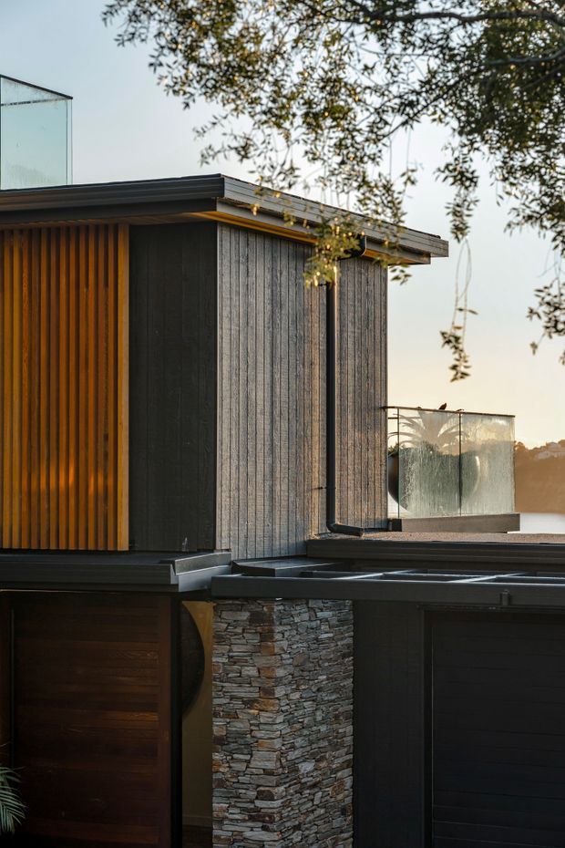 A close exterior view of a contemporary multi-level residence, showing dark vertical timber cladding, warm timber battens, charcoal roof edges and garage doors, a stacked natural stone feature wall, and glass balustrades around an elevated terrace. Mature tree branches partially frame the upper portion of the image.