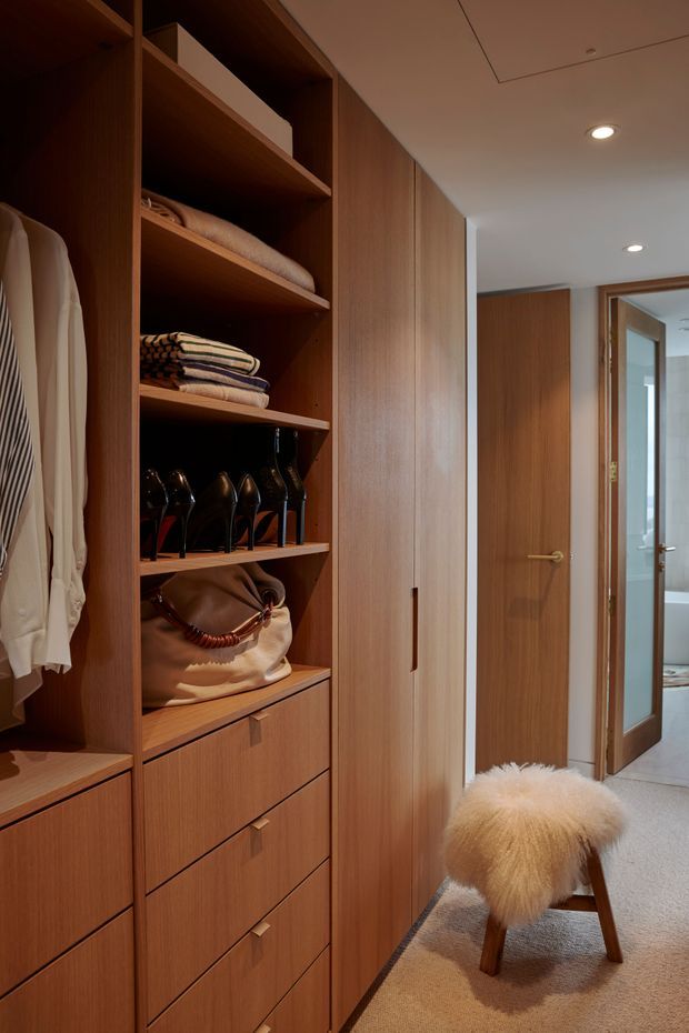 A warm, contemporary walk-in wardrobe with extensive built-in timber joinery. Open shelving displays folded clothing, shoes and a handbag, while drawers and tall cupboards provide concealed storage. A plush fur-covered stool sits on a soft neutral carpet, and a timber door opens toward a bright adjoining bathroom or hallway.