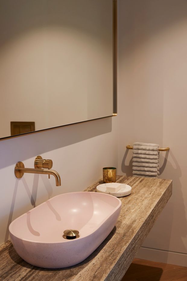 A compact, warmly lit bathroom vanity featuring a freestanding pale pink vessel basin on a thick natural stone countertop. Brushed brass wall-mounted tapware, a large frameless mirror, brass towel rail with folded towels, and a small soap dish complete the refined fit-out. Timber flooring is visible below.