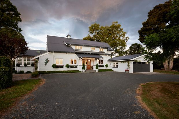 A large two-storey suburban residence viewed from the driveway, with white rendered walls, dark grey pitched roofs, dormer-style upper windows, covered entry, and a detached matching garage. Mature trees and landscaped edges surround the broad paved forecourt.