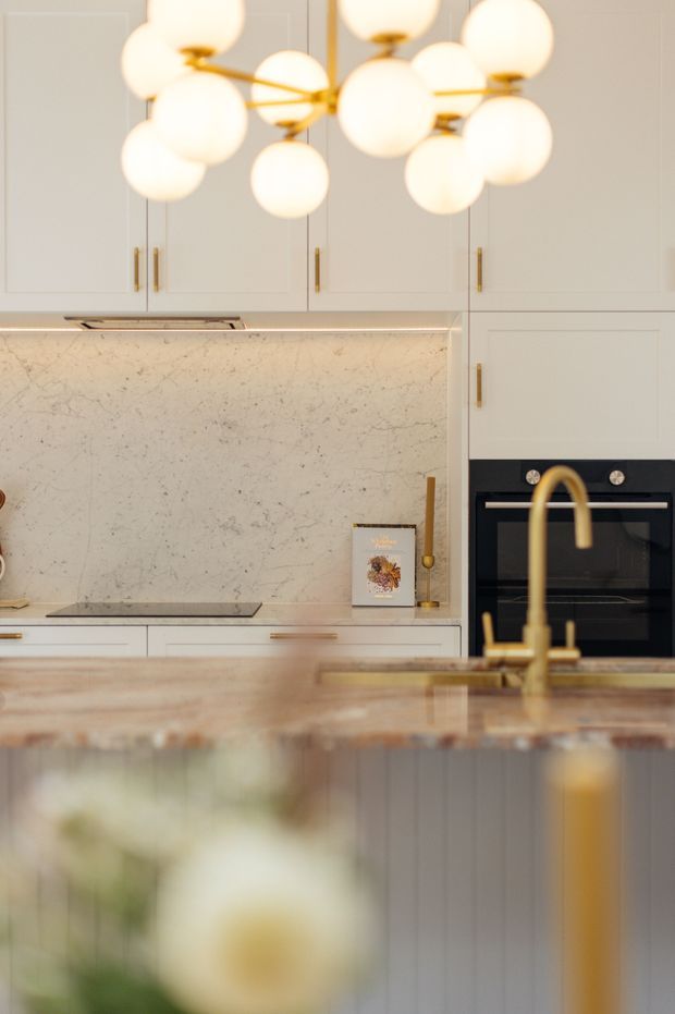 A refined contemporary kitchen viewed across a large island. The kitchen features pale grey-white fixed cabinetry with slim brass-gold handles, a veined light stone splashback and benchtop, an integrated black oven, and a prominent sculptural pendant light with multiple opal globes and brass arms. A brass gooseneck tap rises from the island in the foreground, with softly blurred decorative elements below.