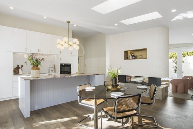 Bright open-plan kitchen and dining area with white shaker-style cabinetry, a large island with fluted detailing, dark stone benchtop, integrated appliances, and a round glass dining table with woven chairs. A curved plaster fireplace wall with recessed display niche separates the kitchen from the living area. Skylights, recessed lighting, pale walls, and timber flooring create an airy, refined contemporary interior.