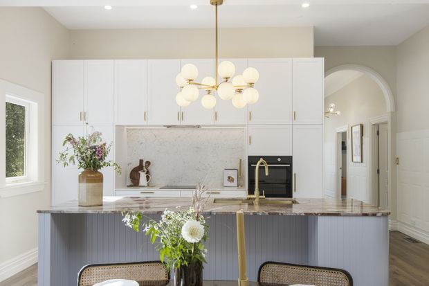 Bright, refined kitchen with a large central island, white full-height cabinetry, pale marble-look splashback and benchtops, integrated black oven, and a fluted grey island base. A sculptural pendant light hangs above the island, with floral arrangements and dining stools adding softness. The room includes traditional architectural detailing such as white wall panelling, cornices, and a large arched opening leading to a hallway.
