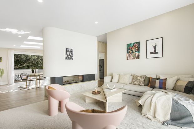 Bright, open-plan contemporary living room with a large pale sectional sofa, sculptural pink lounge chairs, a geometric light-toned coffee table, and a central white fireplace feature wall. The room opens to a dining area and garden-facing glazing, with recessed ceiling lighting, neutral finishes, and colourful artwork.