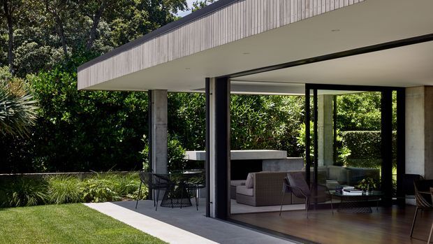 Modern single-storey home exterior with a broad flat roof, exposed concrete columns, extensive black-framed floor-to-ceiling glazing, and a covered outdoor terrace opening onto a landscaped garden. The interior living area and timber flooring are visible through the glazing.